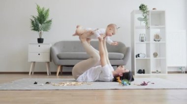 Güzel esmer kadın, oturma odasında halıda uzanırken kucağında kolları ve bacaklarıyla küçük bir kız çocuğu tutuyor. Sevgi dolu anne, küçük kızının eğlenceli oyunlarında güvenle uçmasına izin veriyor..