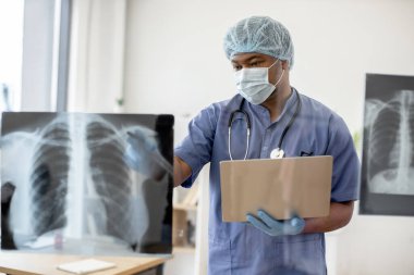 Doktor önlüğü, maske ve tıbbi başlık takmış, doktorların işyerindeki cam duvardaki röntgen taramalarını inceleyen düşünceli Afro-Amerikan bir adam. Hasta hakkında bilgi toplarken dizüstü bilgisayar kullanan pratisyen..