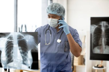 Mavi önlüklü ve yüz maskeli düşünceli bir yetişkin, profesyonel muayenehanede röntgen görüntülerini tararken telefonla konuşuyor. Terapist hastayla sağlık durumunu tartışıyor.