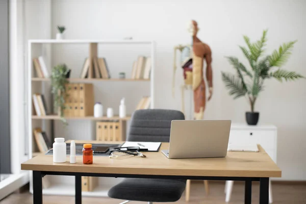 stock image Focus on writing desk with modern laptop, stethoscope, and medicine bottles against background of human skeleton model at doctors office. Spacious consulting room in outpatient department.