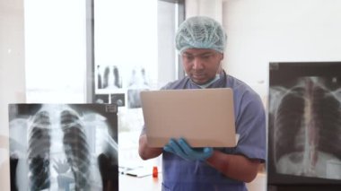 Tıbbi önlüklü, kep ve maske takan, cam panelin önünde göğüs taraması yapılan çok ırklı bir erkek. Hastanede tıbbi hastalık teşhisi koyan deneyimli bir doktor..