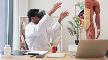 Laboratuvar önlüğü ve 3D gözlük takan Afro-Amerikan adam modern cihazlar ve insan iskeletinin yanında otururken VR sistemi kullanıyor. Eğitim hastanesinde tıbbi eğitime hazırlanan bir terapist..