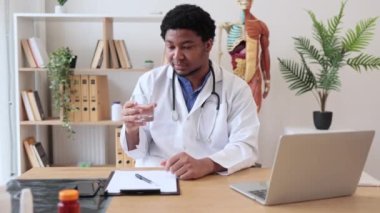 Laboratuvar önlüklü pozitif çok kültürlü doktor dizüstü bilgisayar ve dokümanlarla yazı masasında otururken bir bardak su gösteriyor. Pratisyen hekime susuz kalmamasını önerdim..