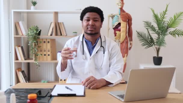 Pozytywny Wielokulturowy Lekarz Fartuchu Laboratoryjnym Pokazujący Szklankę Wody Siedzący Przy — Wideo stockowe