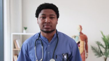 Arka planda insan anatomisi modeliyle doktor ofisinde duran mavi önlüklü ve steteskoplu çekici çok kültürlü adamın portresi. Tıp merkezinde poz veren neşeli bir cerrah..