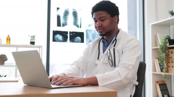 有魅力的非洲裔美国男性穿着实验室外套在笔记本电脑上工作 坐在诊所会客室的办公桌前 在舒适的工作场所研究网上疾病的全科医生 — 图库视频影像