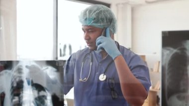 Mavi önlüklü ve eldivenli düşünceli çok kültürlü bir erkek klinikte içerdeyken telefonla konuşuyor. Tıbbi uzman, ilk teşhisi tedavi merkezine açıkladı..