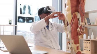 Beyaz önlüklü çok ırklı bir erkeğin ofis masasında anatomik modelin yanında dijital aletlerle otururken sanal gerçeklik kulaklığı kullanması. Profesör klinikteki tıbbi durumu taklit ediyor..
