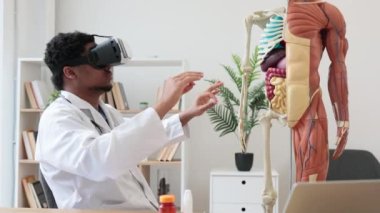 Beyaz önlüklü çok ırklı bir erkeğin ofis masasında anatomik modelin yanında dijital aletlerle otururken sanal gerçeklik kulaklığı kullanması. Profesör klinikteki tıbbi durumu taklit ediyor..