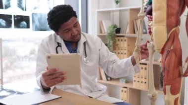 Laboratuar önlüklü, kendinden emin, çok kültürlü bir yetişkin. Doktorun ofisinde elinde tablet tutarken insan anatomisi modelinin uyluk kemiğini gösteriyor. Doktor hasta eğitimi için bilgi topluyor.