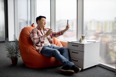 Günlük kıyafetlerini giymiş neşeli Asyalı adam akıllı telefondan video çağrı uygulaması üzerinden çevrimiçi konferans düzenliyor. Kahkaha amiri iş arkadaşlarıyla anahtar sorular hakkında Webinar 'ı tutuyor.