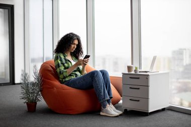 Güzel Afro-Amerikalı kadın günlük kıyafetleriyle cep telefonundan mesaj yollarken dosya depolama yerindeki fasulye çuvalında oturuyor. Yaratıcı iş yerinde telefon uygulaması üzerinden e-posta yazan düşünceli serbest çalışan.
