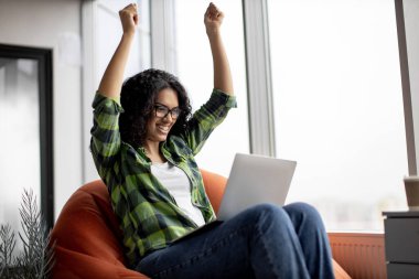 Heyecanlı Afro-Amerikan kadın günlük kıyafetleri içinde dizüstü bilgisayar ekranına bakarken heyecan içinde yumruklarını kaldırıyor. Görevde e-posta okurken kariyer başarısını kutlayan başarılı lider.