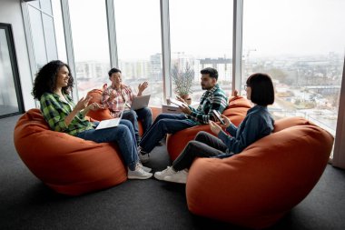 Bir grup genç iş adamı, iş yerinde terracotta puf minderleri üzerinde otururken gündelik giysiler içinde modern aletler taşıyorlar. Yeni proje hakkında bilgi arayan çok kültürlü takım arkadaşları.