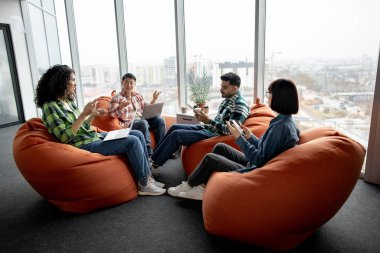 Dört etnik çeşitlilikte meslektaştan oluşan genç bir ekip açık ofiste puf koltuklarda otururken sohbet ediyorlar. Gündelik çalışan aktif iş arkadaşları dijital cihazlar kullanarak iş konularını tartışıyor..