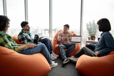 Çeşitli iş adamları modern aletlerle terracotta armut çantalarında otururken Asyalı adam çalışma alanında dizüstü bilgisayar gösteriyor. Veri analistleri proje istatistikleri hakkında beyin fırtınası yapıyorlar.