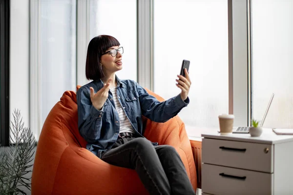 평상복 차림을 안경을 현대식 사무실에서 스마트폰으로 비디오 통화를 모습을 동료들에게 — 스톡 사진