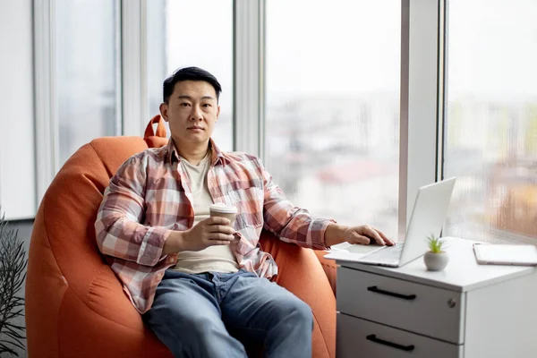 Motivated asian employee sitting on orange comfortable bean bag chair holding cup of aromatic coffee and working on laptop. Young male creating business project using modern wireless technology.