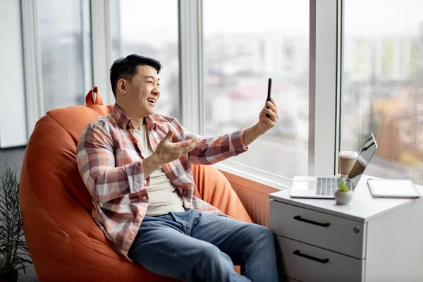 Cheerful asian man dressed in everyday clothes conducting online conference over video call application on smartphone. Laughing supervisor holding webinar about key questions with colleagues.