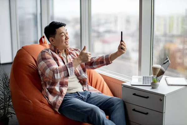 Cheerful asian man dressed in everyday clothes conducting online conference over video call application on smartphone. Laughing supervisor holding webinar about key questions with colleagues.