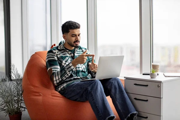 Handsome indian man in casual attire speaking on video call on laptop and gesturing hands in cozy office. Young bearded employee sitting in white spacious workplace with great panoramic view on city.
