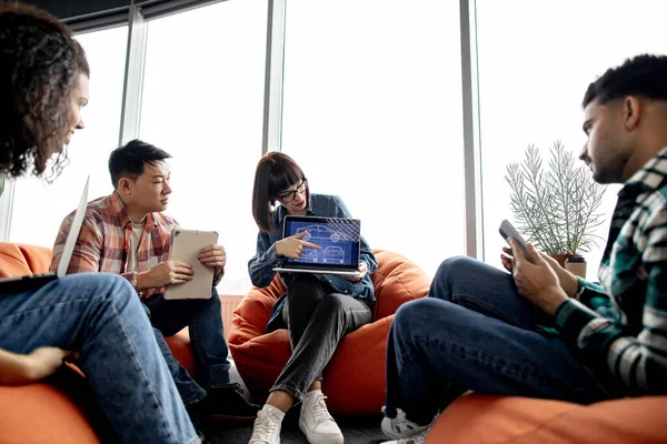 Beautiful caucasian lady in street style clothes pointing at laptop screen attracting attention of international colleagues in office. Car designers participating in auto development process at work.