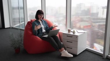 Kafkasyalı beyaz adamın uzaktan görüntülü konuşması ve çalışma süreciyle ilgili önemli sorular hakkında ekiple konuşması. Koyu renk saçlı, gözlüklü, el kol hareketi yapan dizlerinde dizleri olan bir kadın..