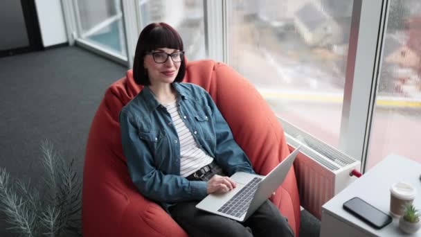 白种人女士 身穿斜纹棉布夹克和黑色牛仔裤 坐在椅子上 对着相机微笑 有魅力的女性在现代合作空间使用无线笔记本电脑进行远程工作 — 图库视频影像