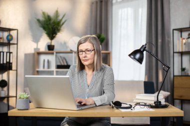 Orta yaşlı, tasmasız bluzuyla gülümseyen, ofisteki yazı masasında otururken taşınabilir bilgisayarda yazan bir kadın. Gözlüklü çekici iş kadını uzaktan elektronik cihaz kullanıyor..