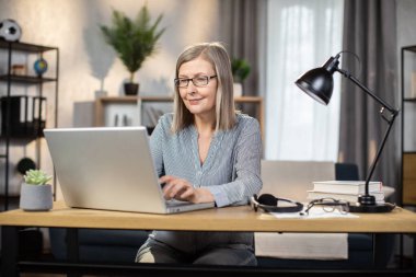 Orta yaşlı, tasmasız bluzuyla gülümseyen, ofisteki yazı masasında otururken taşınabilir bilgisayarda yazan bir kadın. Gözlüklü çekici iş kadını uzaktan elektronik cihaz kullanıyor..