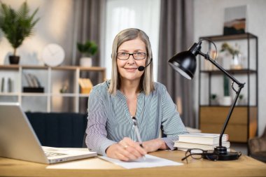 Gözlüklü ve kulaklık takan kendine güvenen iş kadınının portresi bulanık oda arka planında elinde kalem tutuyordu. Modern dijital cihazlar kullanarak evden uzakta çalışan neşeli yaşlı kadın..