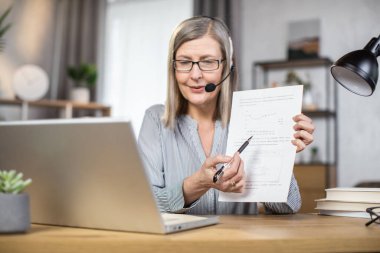 Uzak bir iş yerinde online konferansa ev sahipliği yaparken elinde kalemle kağıt tutarak ciddi bir orta yaşlı kadın. Olgun baş yönetici, bilgisayar kamerasıyla proje verilerini meslektaşlarıyla tartışıyor.