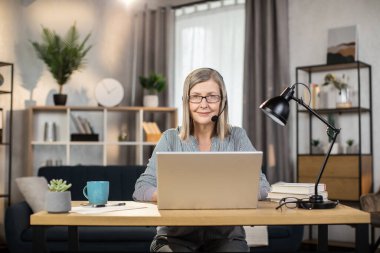 Evdeki geniş iş yerinde dizüstü bilgisayarın arkasında otururken, elsiz kulaklıklı gülümseyen kadın işçinin portresi. Çevrimiçi takım arkadaşlarıyla dikkatli bir çalışanın grup toplantısı.