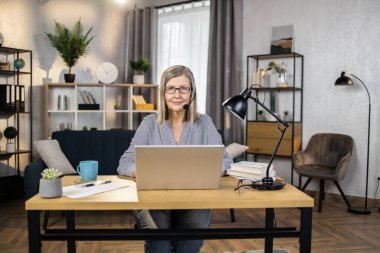 Evdeki geniş iş yerinde dizüstü bilgisayarın arkasında otururken, elsiz kulaklıklı gülümseyen kadın işçinin portresi. Çevrimiçi takım arkadaşlarıyla dikkatli bir çalışanın grup toplantısı.