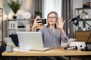 Uzak ofisteki webcam 'e el sallarken elinde cep telefonu tutan orta yaşlı çekici beyaz bir bayan. Dijital aygıt aracılığıyla video çağrısı alırken neşeli yönetici meslektaşlarını selamlıyor.