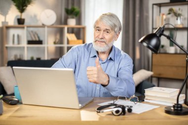 Olgun bir iş adamı, uzak bir ofiste dizüstü bilgisayar kullanırken başparmak hareketi yapıyor. Düğmeli gömlekli rahat serbest çalışan ev içindeki iş-yaşam dengesini onayladığını ifade ediyor..