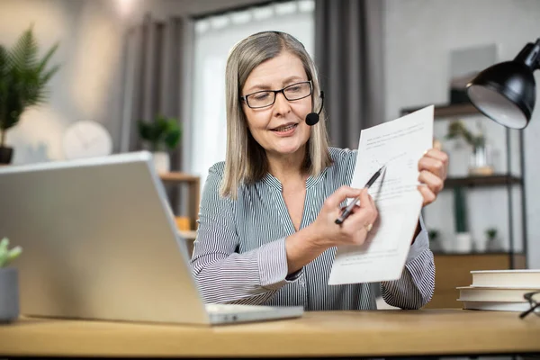 Focus on elderly lady in formal wear and glasses showing documents while sitting with headset at desk in home office. Elegant entrepreneur conducting online briefing with company staff via webcam.
