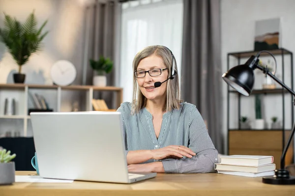 Portrait of smiling female worker in hands-free headset sitting behind laptop at office desk in spacious workplace at home. Mindful remote employee scheduling group meeting with teammates online.