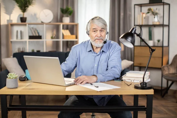 Serious middle-aged male in headset with microphone making video call while sitting at writing desk in home office. Mindful remote employee discussing business issues with coworkers via online app.
