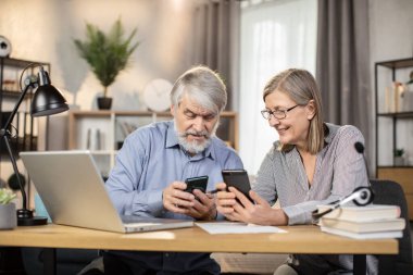 Modern cep telefonlarını tutan mutlu, yaşlı eşler ofis aletleri üzerinde uzak mesafelere dinleniyorlar. Neşeli orta yaşlı serbest çalışanlar kısa molada sosyal medyayı cep telefonlarıyla kontrol ediyorlar.