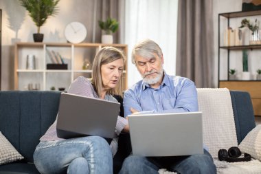Evde rahat bir ortamda vakit geçirirken dizüstü bilgisayar kullanan ciddi yetişkin insanlar. Yaşlı bir aile babası ve karısı kanepede otururken dijital ekranda online makale okuyor..