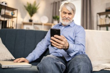 Yaşlı bir erkek tarafından taşınan siyah cep telefonuna odaklan. Diğer elinde bilgisayarla kanepede dinleniyor. Sakin kot pantolon ve düğmeli gömlek giyen yaşlı yetişkin boş zamanlarında online etkinliğe abone oluyor.