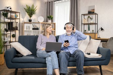 Kablosuz kulaklık ve cep telefonu kullanan neşeli olgun bir adam orta yaşlı güzel bir kadın dinlenme odasında dizüstü bilgisayarla dinlenirken. Üst düzey eşler evlerinde modern müzik listelerinden harika müzikler dinliyorlar..