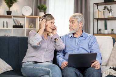 Neşeli son sınıf çifti modern dizüstü bilgisayarlarıyla şık oturma odalarında komedi programı izlemek için rahat koltukta eğleniyorlar. Gülümseyen kadın, sevgili kocası için kablosuz kulaklık ayarlıyor..