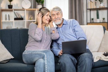 Yaşlı sevimli çift rahat koltukta dinleniyor ve evde romantik müzik dinliyor. Kocası dizlerinin üstünde kişisel laptop tutarken, son sınıf güzel bayanın kablosuz kulaklığı var..