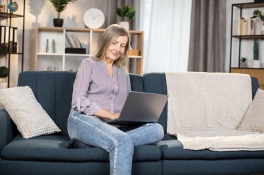 60 'lı yaşlarda beyaz bir kadın taşınabilir laptopuyla yumuşak koltukta dinleniyor ve sosyal medyada geziniyor. Rahat bir evde modern aletler kullanarak gündelik giysiler içinde gülümseyen kadın..