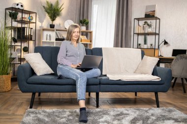 60 'lı yaşlarda beyaz bir kadın taşınabilir laptopuyla yumuşak koltukta dinleniyor ve sosyal medyada geziniyor. Rahat bir evde modern aletler kullanarak gündelik giysiler içinde gülümseyen kadın..