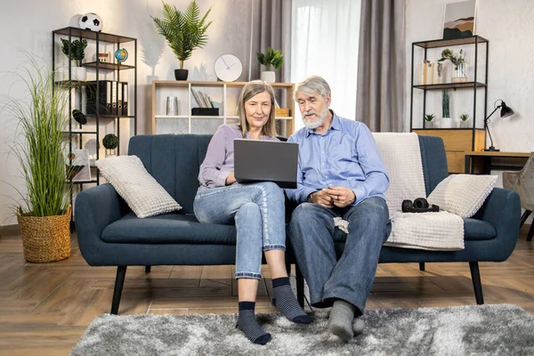 Full length view of relaxed elderly adults watching video on modern laptop while having fun in living room. Happy grandparents relishing kids birthday party videos shared in social media network.