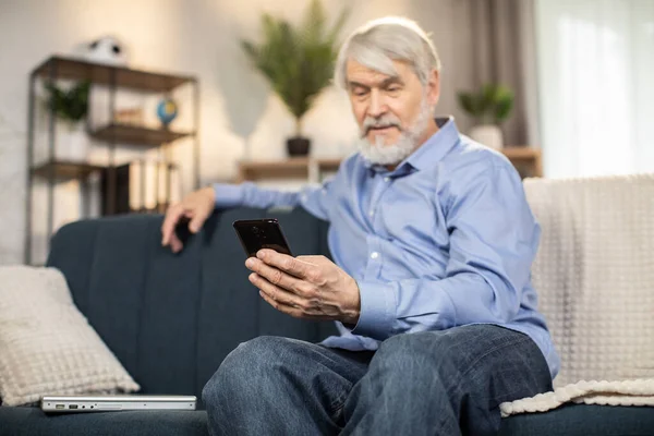室内の居心地の良いソファで休んでカジュアルな服装で高齢者の髭の男が保持する携帯電話に選択的な焦点を当てます 家庭で利用可能なアプリケーションを介して孫に現代の祖父のテキストメッセージ — ストック写真
