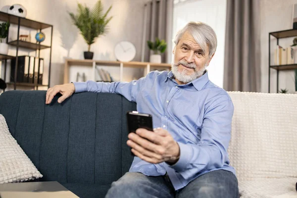 リビングルームの暗いソファに座っている間 スマートフォンでポーズを取るカジュアルな服でシニア白人男性に焦点を当てます 通話やメッセージにモバイル接続を使用してリラックスした白髪の男 — ストック写真